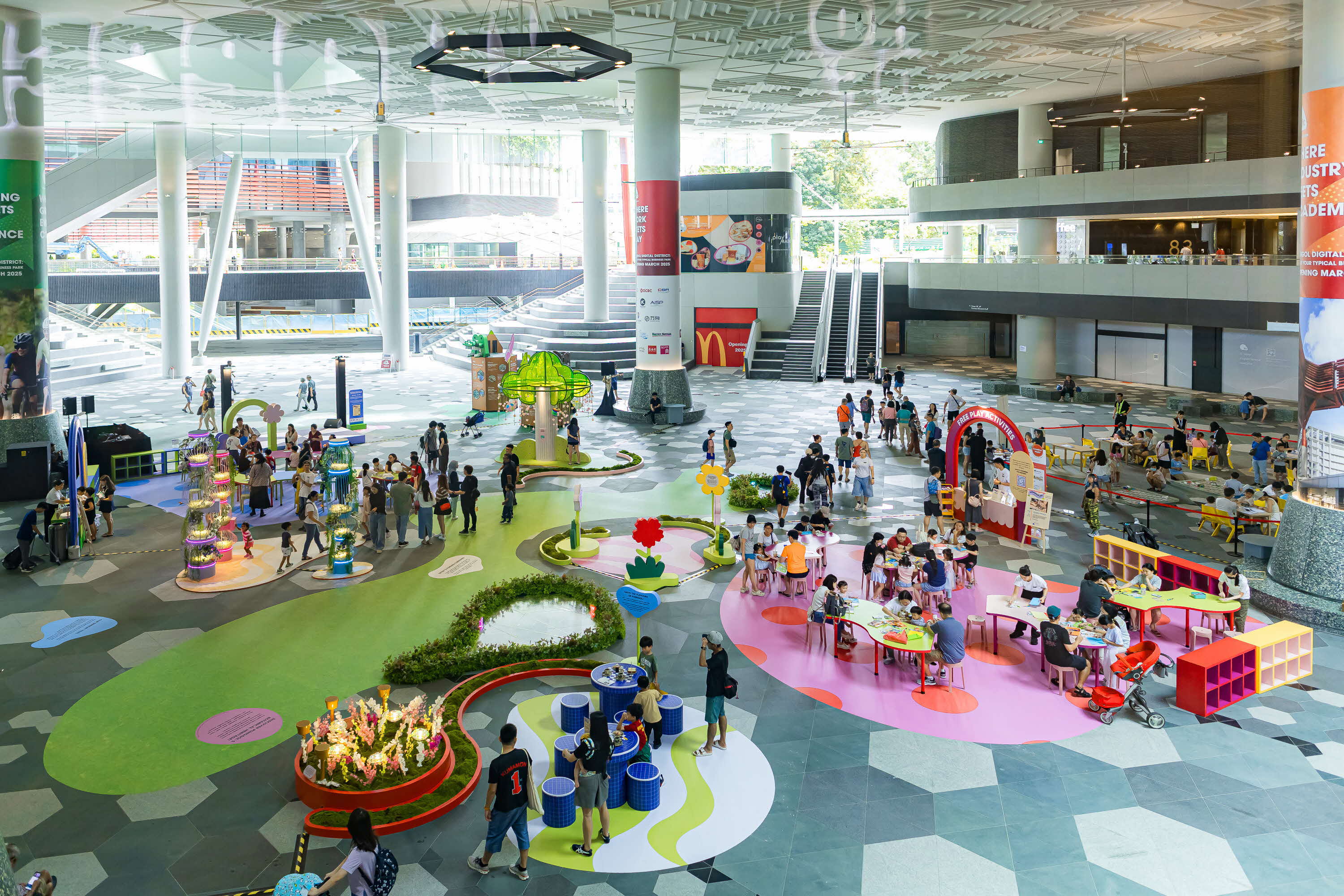An overhead view of an indoor play area with colorful tables, people, and play structures. The area is in a large modern building with escalators and advertisements.